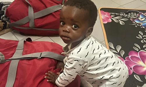 A baby in a white outfit with black patterns stands beside red bags, looking back curiously.