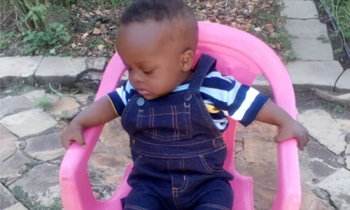 A toddler sitting in a pink chair, wearing a striped shirt and overalls, with a serious expression.