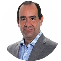 A man with short dark hair wearing a gray suit and blue patterned shirt, smiling against a white background.