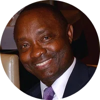 Smiling man in a suit with a purple tie, seated at a dining table with blurred background.