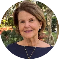 Woman with shoulder-length brown hair and a friendly smile, wearing a necklace, outdoors with greenery in the background.