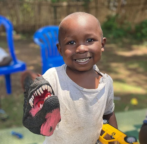 Child smiling while wearing a dinosaur hand puppet, playing outdoors with blue chairs in the background.