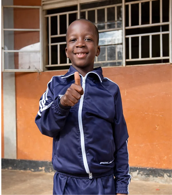 Smiling boy in a blue tracksuit giving a thumbs up in front of a brick wall and window.