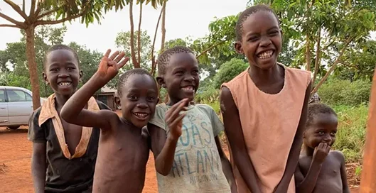Five children smiling and waving in a rural setting with greenery and a dirt path.