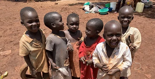 A group of five smiling children in tattered clothing stand together in a dry, rural area.
