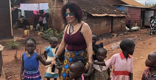Woman wearing sunglasses and colorful clothing walks with children in a village setting.
