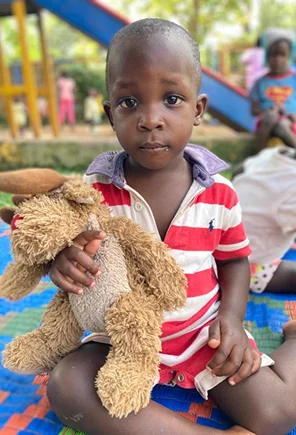 Young child sitting on a colorful mat, holding a stuffed animal, with playground equipment in the background.