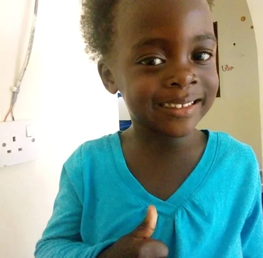 Smiling child in a blue shirt giving a thumbs up, standing against a light-colored wall.