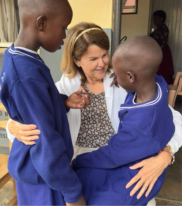 A woman interacts with two children in blue school uniforms, all smiling and engaged in a warm moment together.