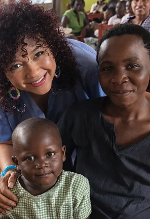 Three people smiling together: a woman with curly hair and earrings, a young child in a green checkered shirt, and a woman.