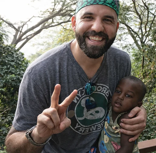 Smiling man with a beard and green bandana holds a young boy, both posing with peace signs amidst greenery.
