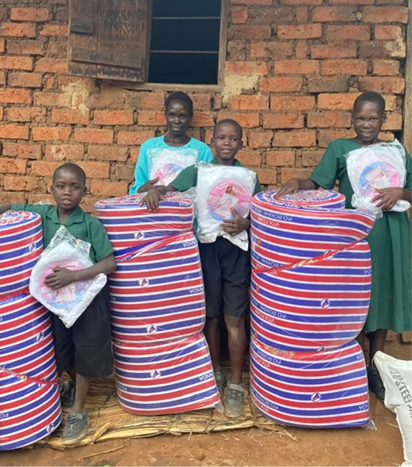 Four children stand in front of a brick wall, holding rolled foam mattresses wrapped in plastic.