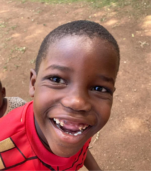 Smiling young boy in a red shirt with a gap-toothed grin, set against a brown earth background.