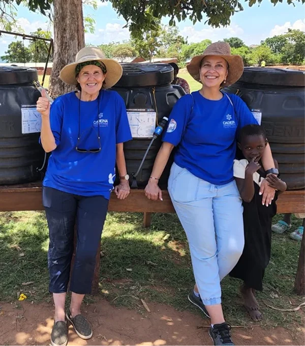 Two women in blue shirts and hats standing beside water storage barrels, smiling, with a child nearby.