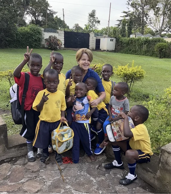 A woman stands with a group of smiling children in yellow uniforms, holding bags, in a green garden setting.