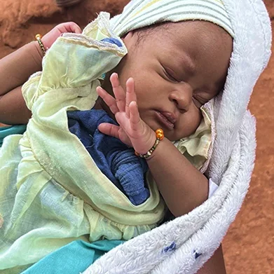 A sleeping baby wrapped in a blanket, wearing a light-colored dress and headscarf, with a small bracelet on their wrist.