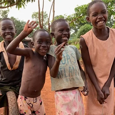Four children smiling and waving, dressed in casual clothing, standing outdoors on red soil with lush greenery in the