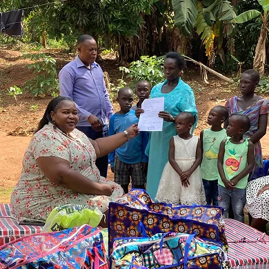 A woman presents a document to a group of children and adults outdoors, surrounded by colorful bags and greenery.