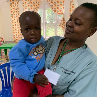 A woman holds a small child, both looking at the camera, in a brightly colored room with children's decor.