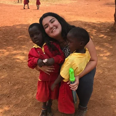 A young woman smiles while hugging two children in colorful clothing on a dirt field.