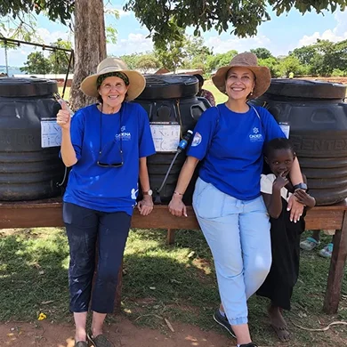 Two women in blue t-shirts and hats stand smiling by large water containers, with a child peeking from behind.