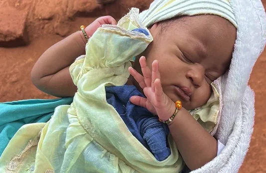 A sleeping baby wrapped in a green outfit, resting with one hand raised, on a sandy surface.