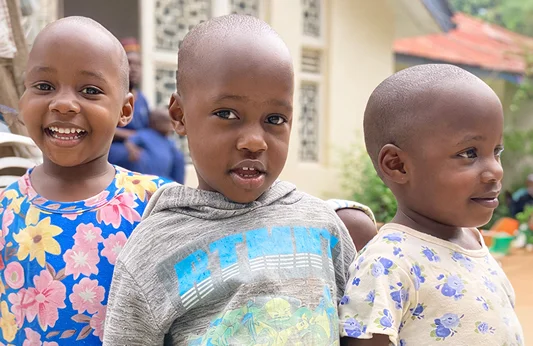 Three smiling children with shaved heads, standing close together in a vibrant, colorful outdoor setting.