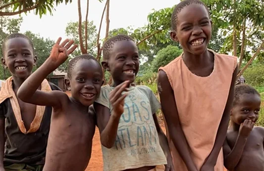 Five smiling children pose together outdoors with trees in the background, some waving and expressing joy.