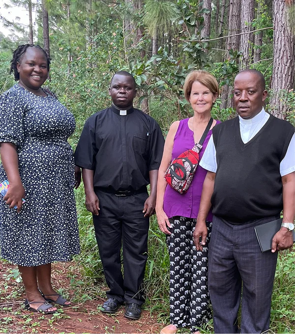 Four individuals stand together in a forested area, surrounded by greenery, smiling at the camera.