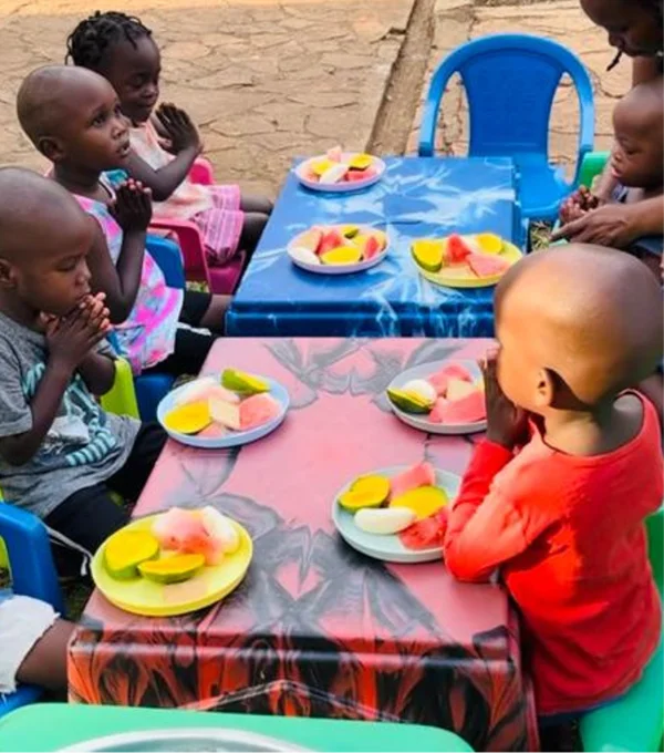 Children sitting at tables, hands clasped in prayer, with plates of fruit in front of them.