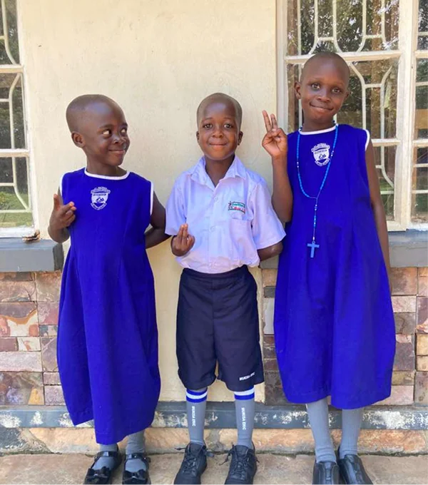 Three children in school uniforms smiling and posing, with one boy making a peace sign outside a building.