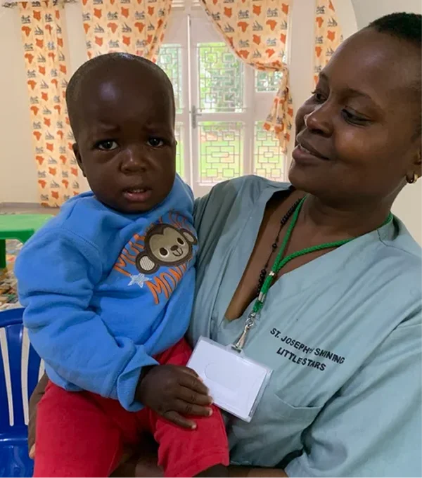 A caregiver holds a small child, both looking at the camera in a bright, cheerful room.