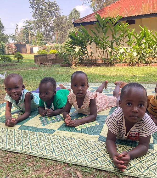 Four children lying on a green mat outdoors, smiling and playing in a sunny garden.