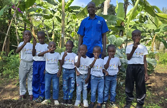 A group of children and an adult pose together in a lush garden, all wearing matching t-shirts.
