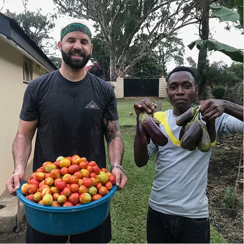 Two men hold fresh produce: one with a blue bucket of tomatoes, the other with eggplants, in a farm setting.