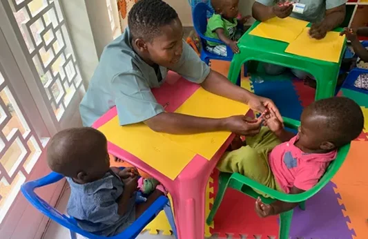 A caregiver interacts with a child at a colorful play area with other children seated nearby.