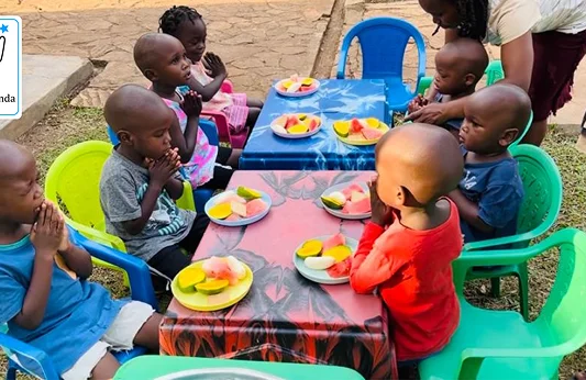 Children sitting at colorful tables with plates of fruit, looking happy while enjoying a meal together outdoors.