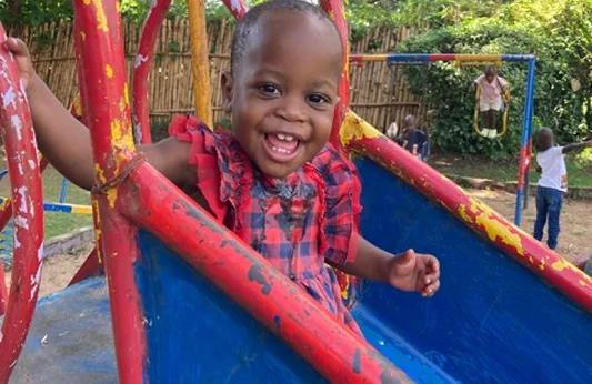 Smiling child in a red-checkered outfit playing on a colorful slide in a playground.