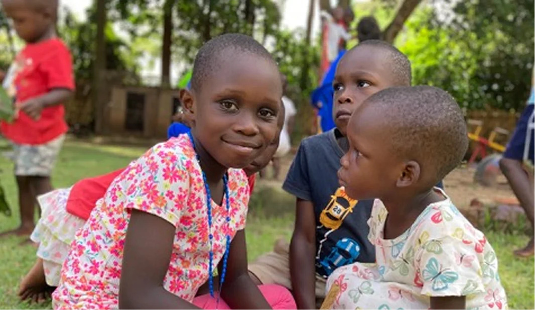 Children playing outdoors; one girl smiles at the camera while two others look on, surrounded by greenery.