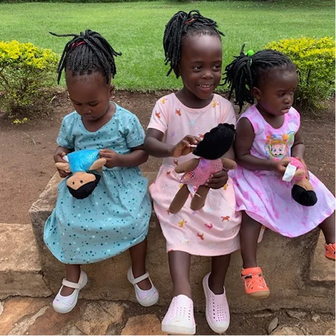 Three children sitting on a stone wall, each holding a doll, wearing colorful dresses in a green outdoor setting.
