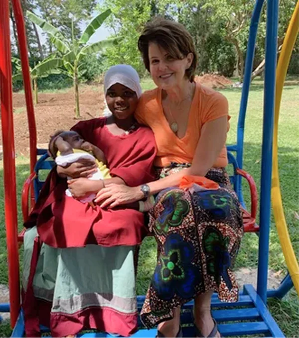 Two women sit together on a swing in a playground, one holding a baby, surrounded by greenery.