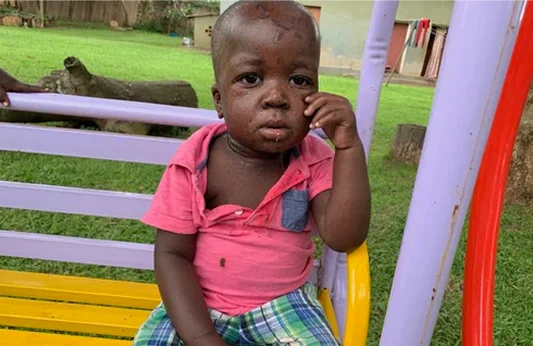 A young child with a focused expression sits on a colorful swing in a grassy playground.