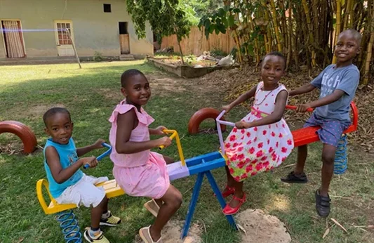 Four children playing on a colorful seesaw in a grassy playground with trees in the background.