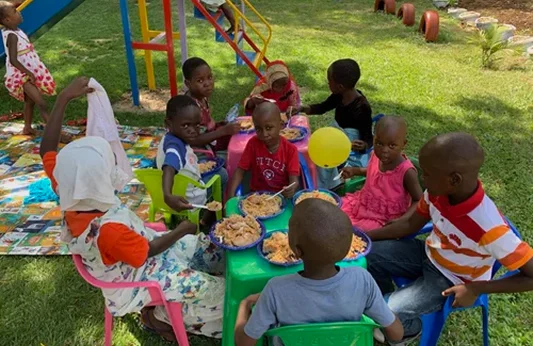 Children sitting around a table in a playground, enjoying food together, with colorful play equipment in the background.