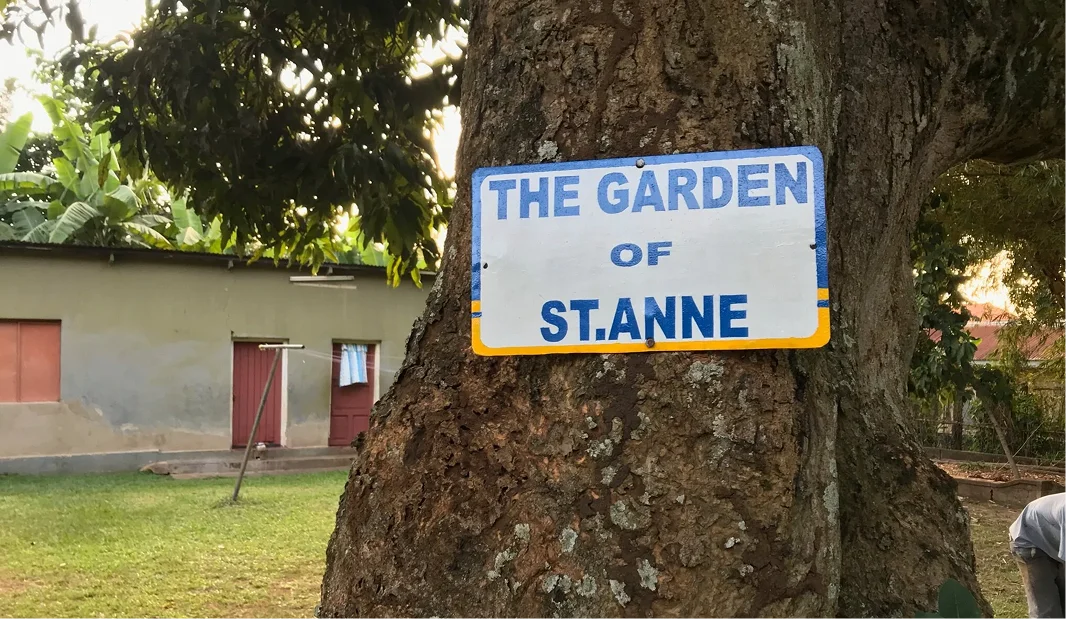 Sign reading "The Garden of St. Anne" attached to a tree, with a building and grassy area in the background.