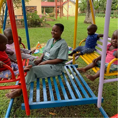 A caregiver sits on a colorful swing surrounded by five children on a playground.