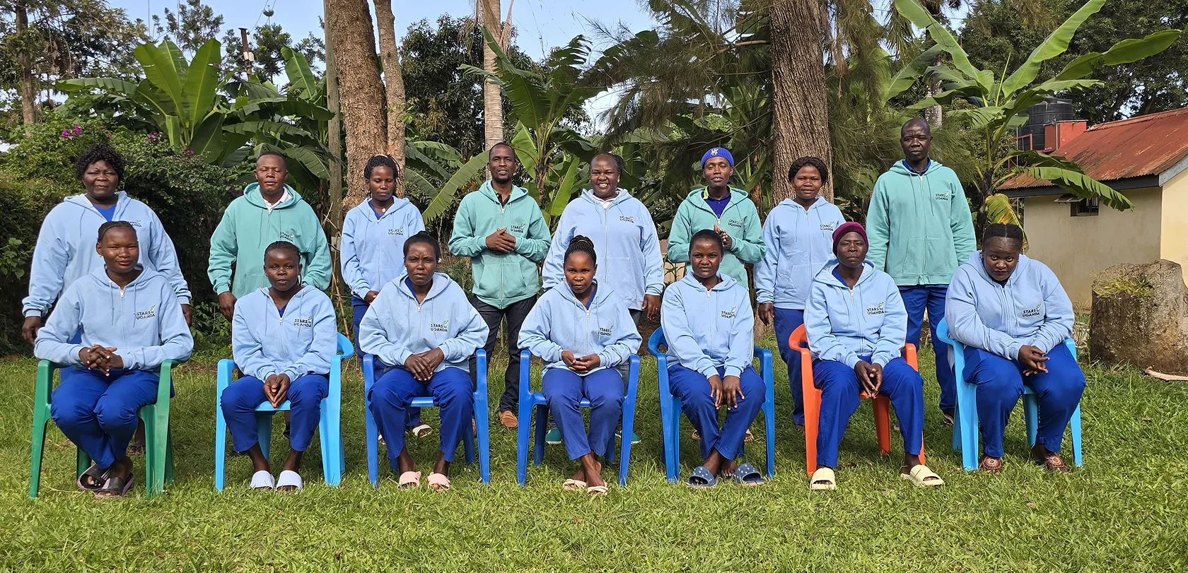A diverse group of 15 individuals in light blue and green attire seated and standing on a grassy area with trees in the