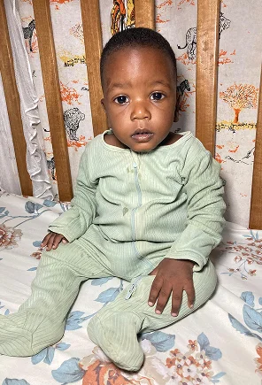 Baby sitting on a crib mattress, wearing a light green onesie, with a curious expression. Background features floral
