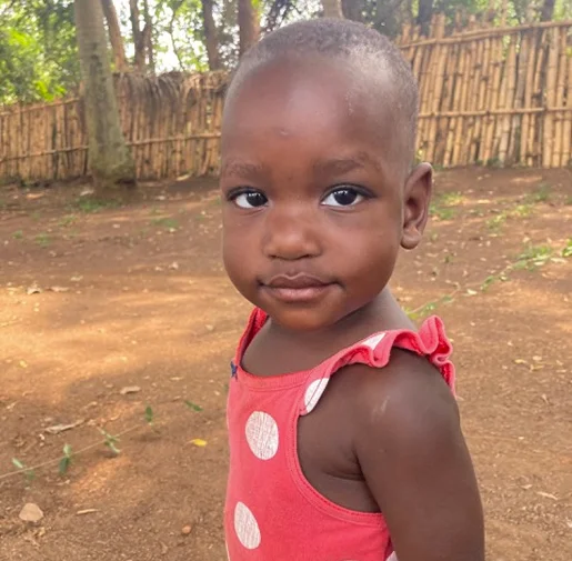 Child in a red dress with white polka dots, looking back over their shoulder, standing outdoors near a bamboo fence.