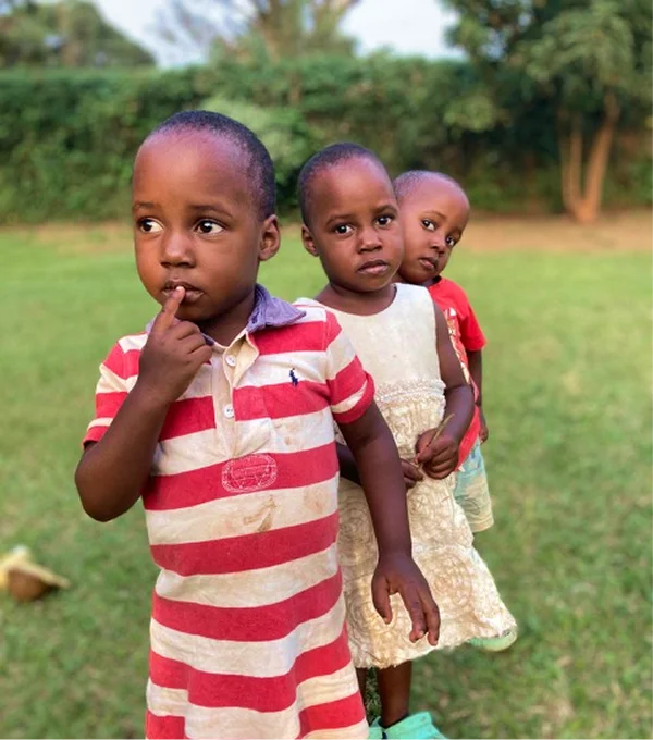Three children stand outdoors in a grassy area, with one child looking thoughtful and the others following closely behind.